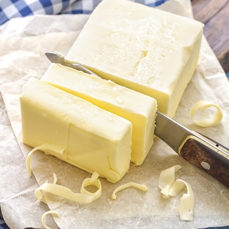 A large cube of butter sits on wax paper, and has two slabs of butter cut off of it with a small paring knife.