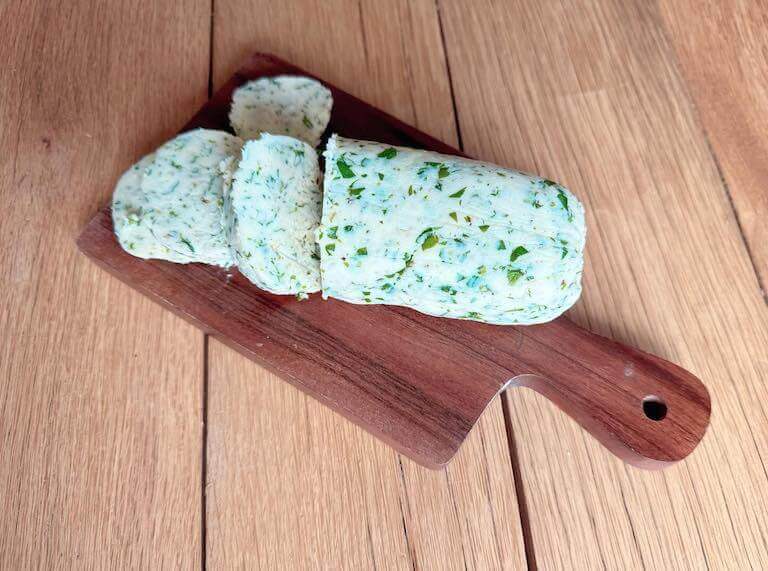 A partially sliced cylinder of butter compounded with green herbs sits on a small wooden board.