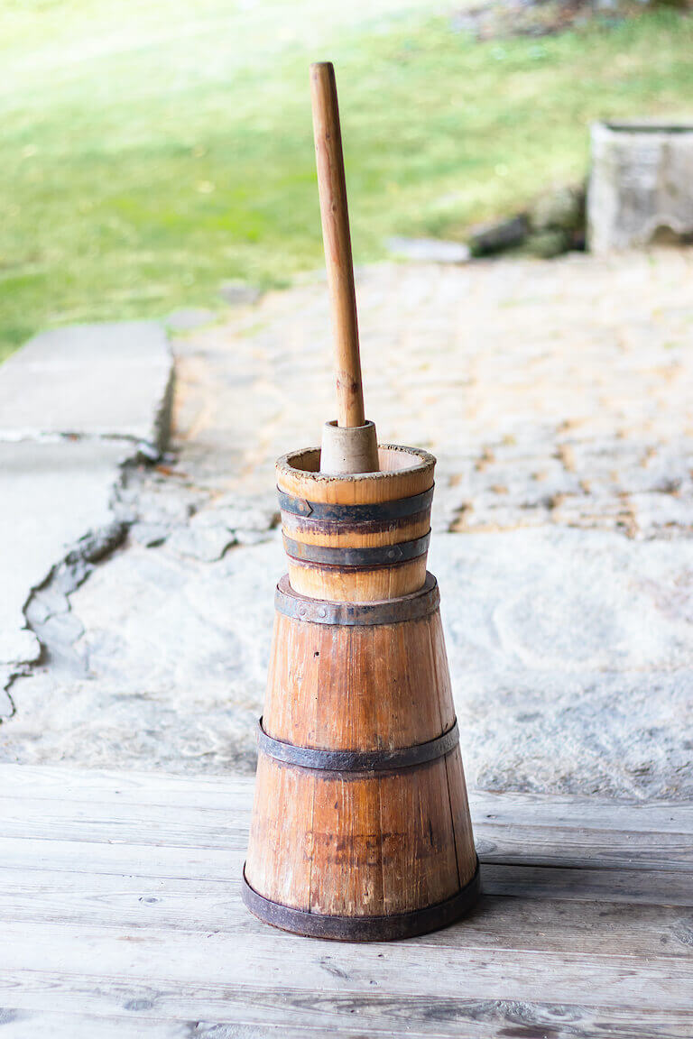 A traditional butter churn is shown. It has a narrow wooden barrel with a paddle handle sticking out the top.