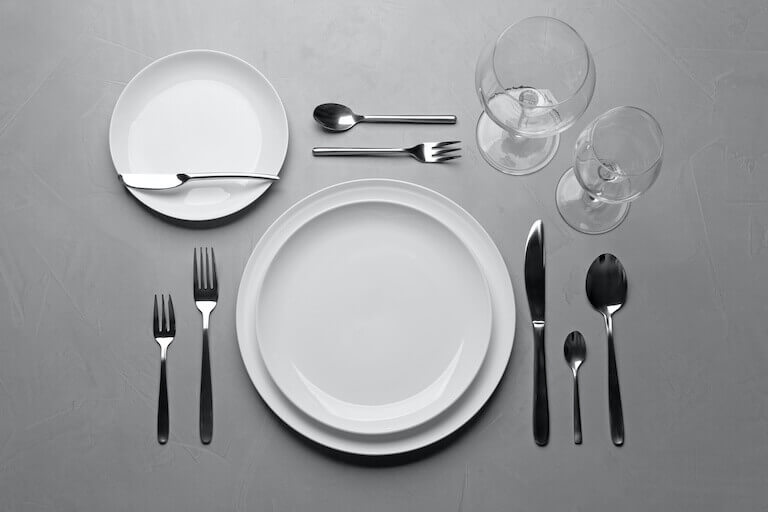 A top-down image of a formal place setting with several glasses, dishes, and pieces of cutlery against a gray surface.
