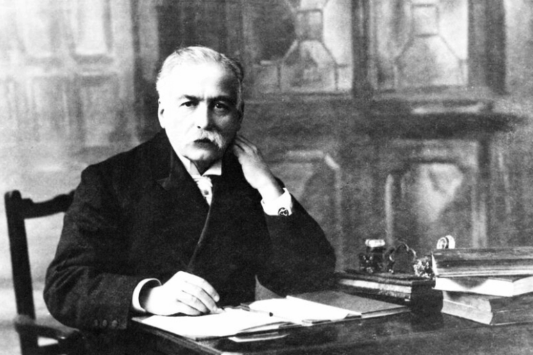 Black and white image of Auguste Escoffier seated at a table in an ornate room, looking up from writing in a notebook.