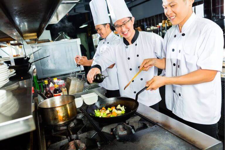 A chef pouring oil in a pan while another chef stirs the vegetables in the pan over the stovetop.