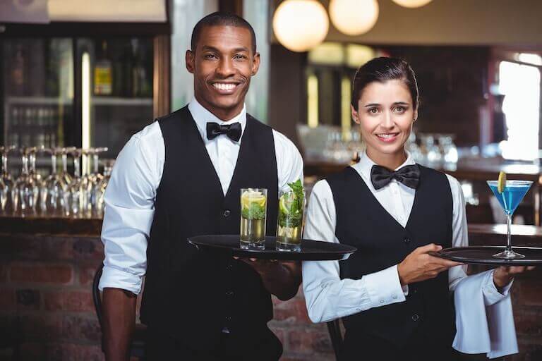 Two servers, each holding serving trays and wearing white button-down shirts with black bow ties, black vests, and black dress pants.