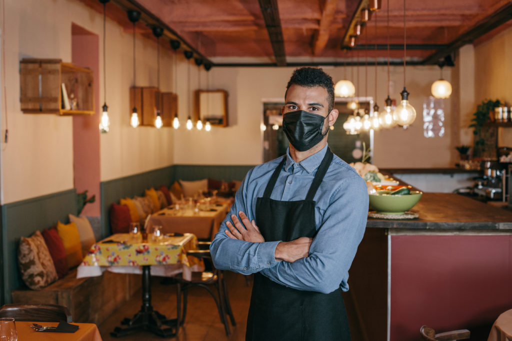  Uomo d'affari afroamericano che indossa una maschera protettiva nera e in piedi al suo ristorante durante il coronavirus.