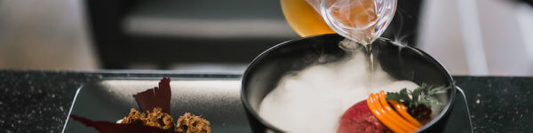 A hand pours an amber liquid over a black bowl containing a red sphere of food surrounded by the smoke of dry ice and set up on a black plate.