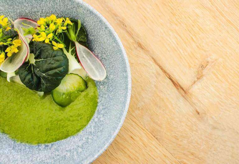 A vibrant green pureed vegetable soup is garnished with kale, broccoli blossoms, and thinly sliced radishes, and displayed in a ceramic bowl.