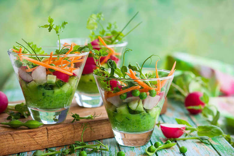 Sliced carrots, peas, and microgreens fill small cone-shaped glasses on an outdoor table.