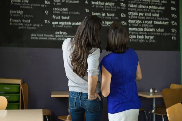 Two women view a restaurant menu on a wall, trying to decide what to order.