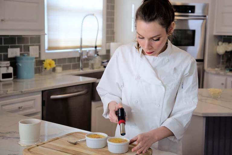 A chef using a blowtorch to caramelize the top of a crème brûlée in a kitchen.