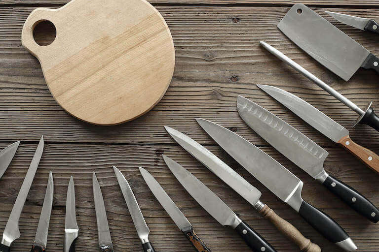 An assortment of various types and sizes of kitchen knives surrounding a circular wooden cutting board on a wooden table