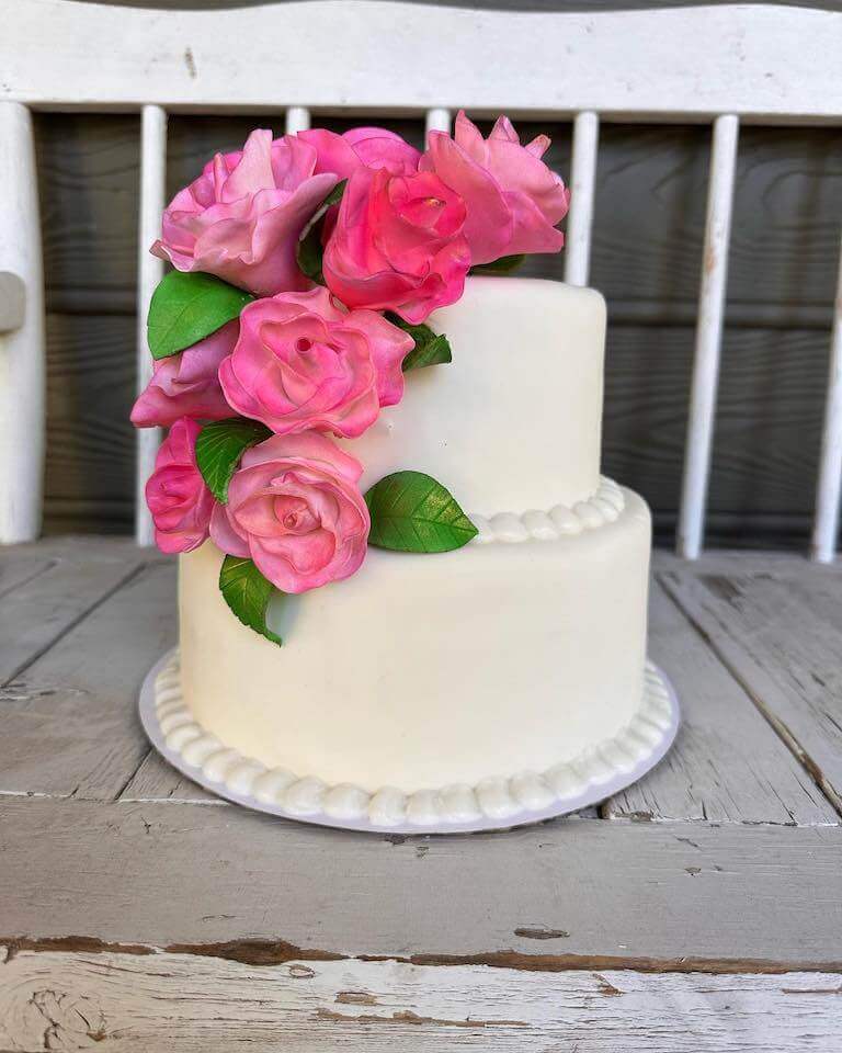 A two-tiered white cake with pink flowers sits outside on a wooden surface.