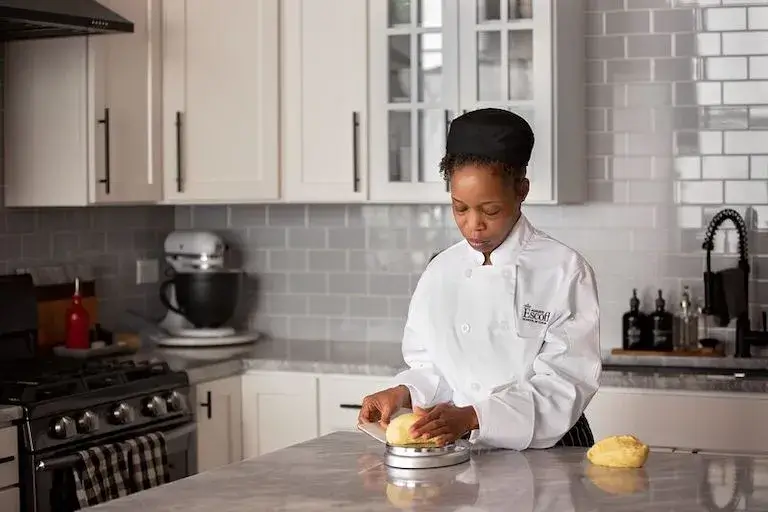 Person in chef jacket using a kitchen scale on the counter to measure dough.