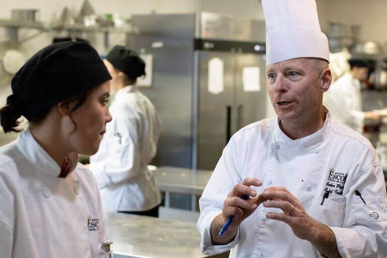 A Chef Instructor stands next to a culinary school student in a commercial training kitchen.