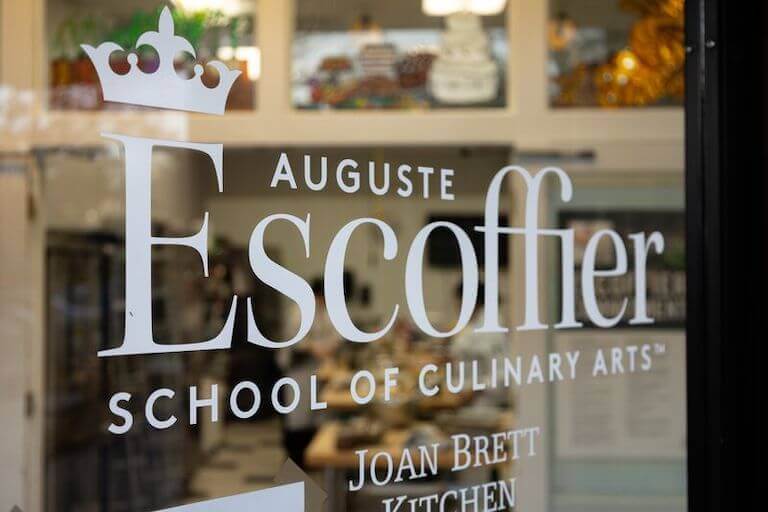 The image shows a close up of the glass front door of the Escoffier Boulder campus, with the words, “Auguste Escoffier School of Culinary Arts” on it.