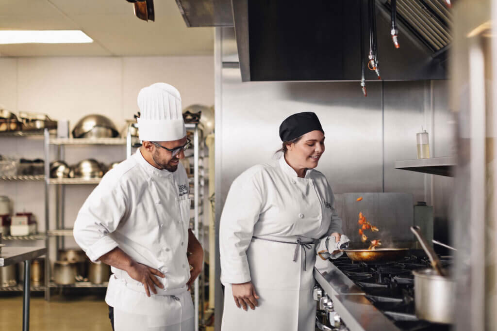 a chef instructor teaching a culinary student over a hot stove