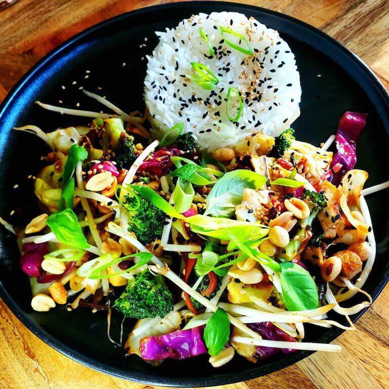 A black plate with sesame rice, fresh sprouts, broccoli, nuts, and other plant-based food. 