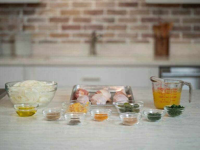 Ingredients for a chicken tagine, such as raw chicken, spices, and oil, are set out in small bowls on a kitchen work surface.