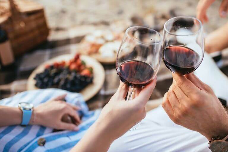 Two people on a picnic toast one another with wine glasses half filled with red wine.