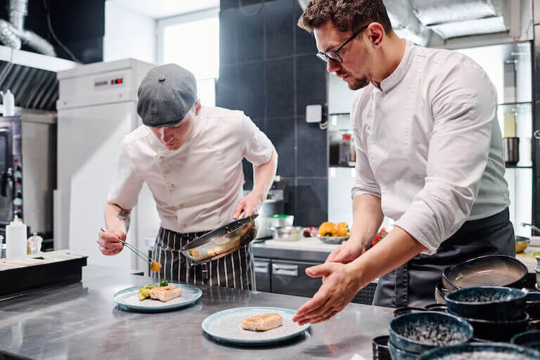 A chef, on the right, shows a cook, on the left, how to properly plate a dish.