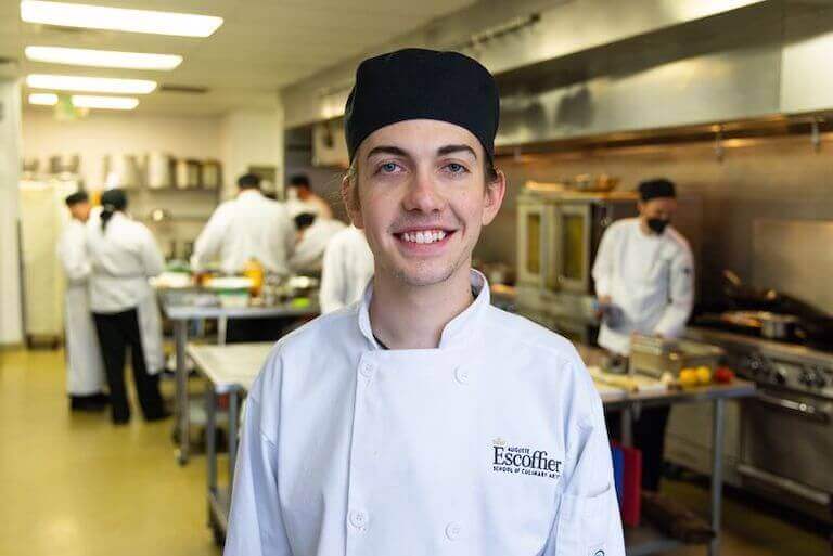 https://www.escoffier.edu/wp-content/uploads/2022/09/Escoffier-student-wearing-their-uniform-in-a-kitchen-smiling-and-posing-for-a-photo.jpeg