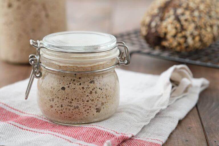 Active rye sourdough starter in a glass jar with a lid on a table with a loaf of bread in the background.