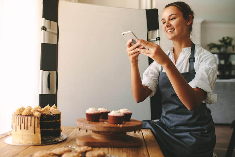 A woman photographs cupcakes and a tiered cake for posting on social media.