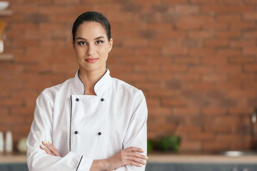 Chef standing in front of a brick in wall in a kitchen with her arms crossed