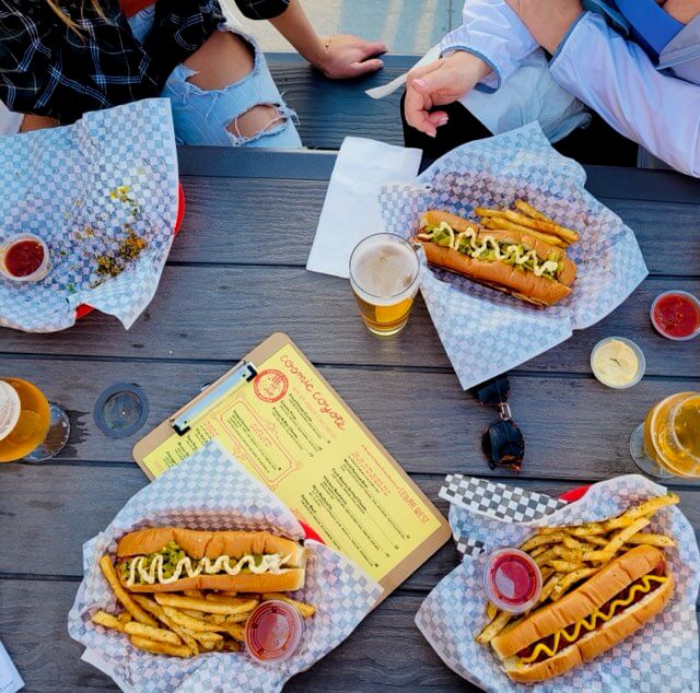 top-down view of hot dogs with all the fixings sittin on a picnic table with menus for Cosmic Coyote