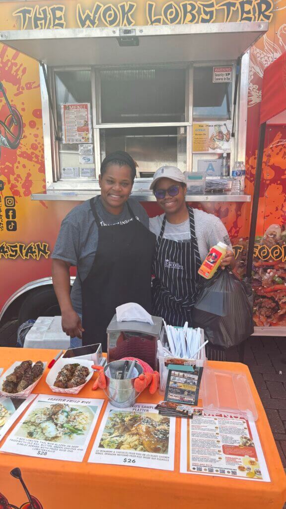 Two cooks stand outside of a food truck behind a table of asian fusion cuisine