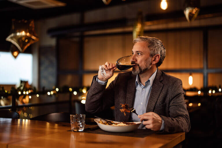 A bearded diner wearing a blazer takes a sip of red wine while seated at a table with a nicely plated entree in a warmly lit restaurant dining room.