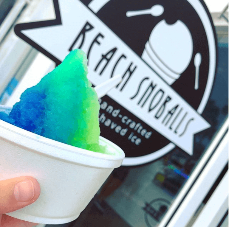 A closeup of a dish of blue and green shaved ice with the Beach Snoballs sign in the background.