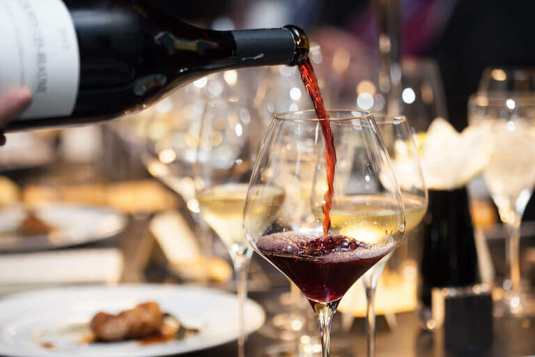 Close-up image of red wine being poured from a bottle into a wine glass, with several other wine glasses in the background.