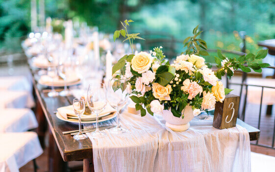 Festive outdoor wedding celebration table covered with natural tablecloth and beautifully, neatly served with sets of dishes, glasses, appliances, and flower arrangements on a green background