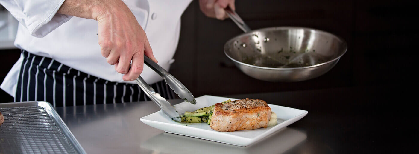 An online culinary arts student plating pork chops and sauteed zucchini on a square white plate in his home kitchen