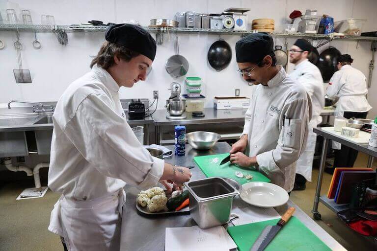 Two people in white culinary uniforms prepare ingredients in a kitchen at Escoffier’s Boulder campus. 