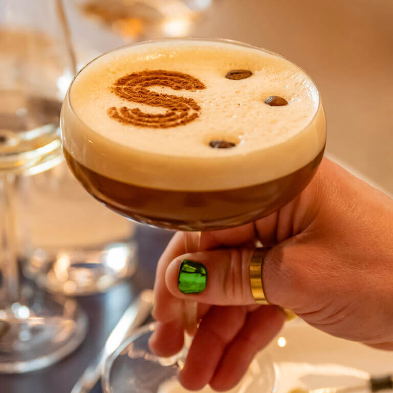 A hand with shiny green nail polish and a gold ring holds an espresso martini in a coupe glass; three espresso beans and the letter ‘S’ stenciled with cocoa powder float on a layer of foam on top of the cocktail.