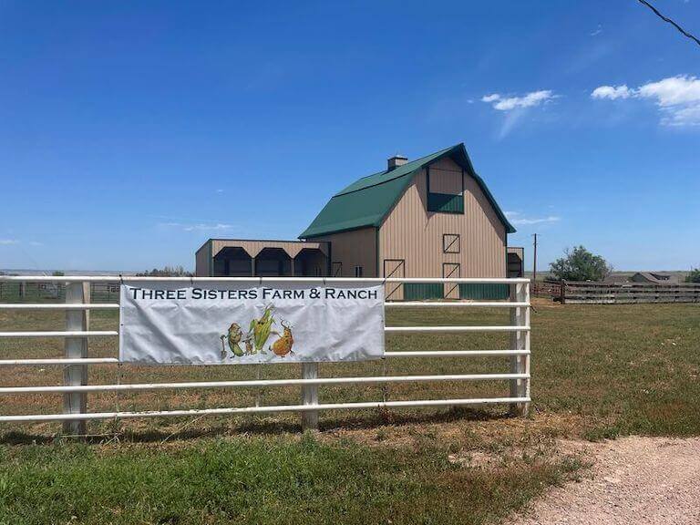 A banner on a ranch gate in front of a large barn reads “Three Sisters Farm.”