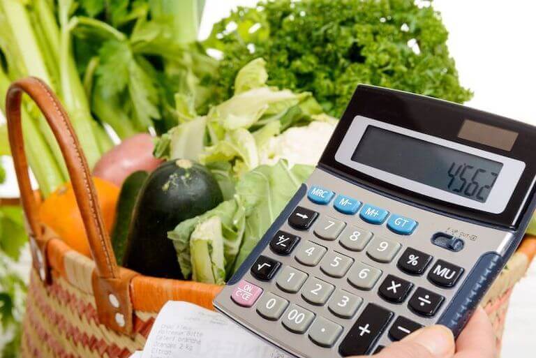 A hand holds a calculator and a basket of vegetables is in the background.