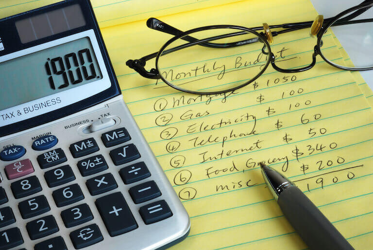 A yellow lined pad of paper with a list of household expenses, with a calculator to the left of the pad and a pair of glasses sitting on top of it.