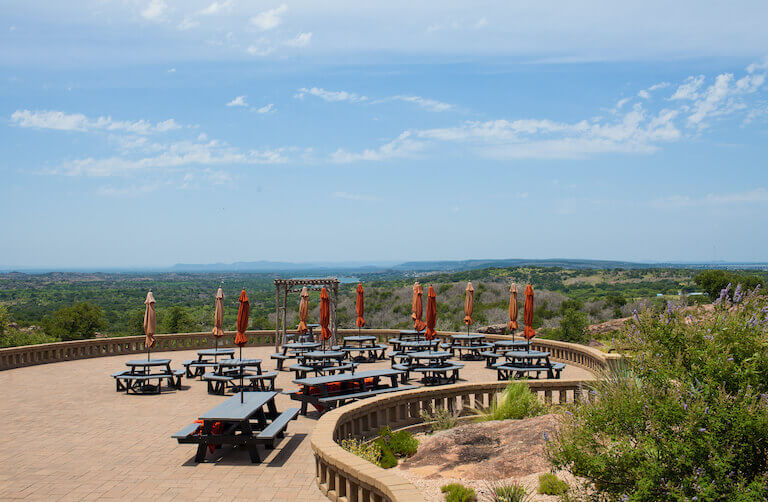 Several picnic tables with umbrellas are spread across a large stone patio overlooking an expansive landscape of lakes and rolling hills under a blue sky. 