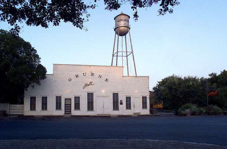 The weathered wooden facade of a low, Western-style building surrounded by trees; an old-fashioned water tower rises up in the background behind the building.