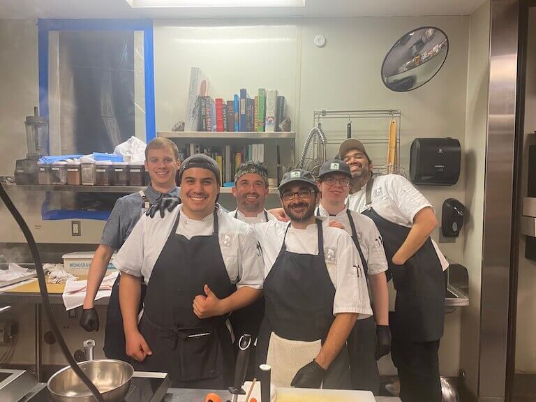 A group of six prep chefs, including Michael Fields, in chef’s whites stand smiling in a commercial kitchen. 