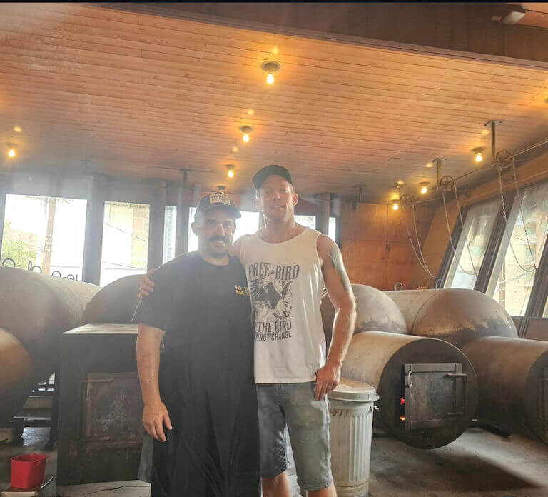 Michael and Aaron Franklin stand in front of four large, cylindrical, metal meat smokers.