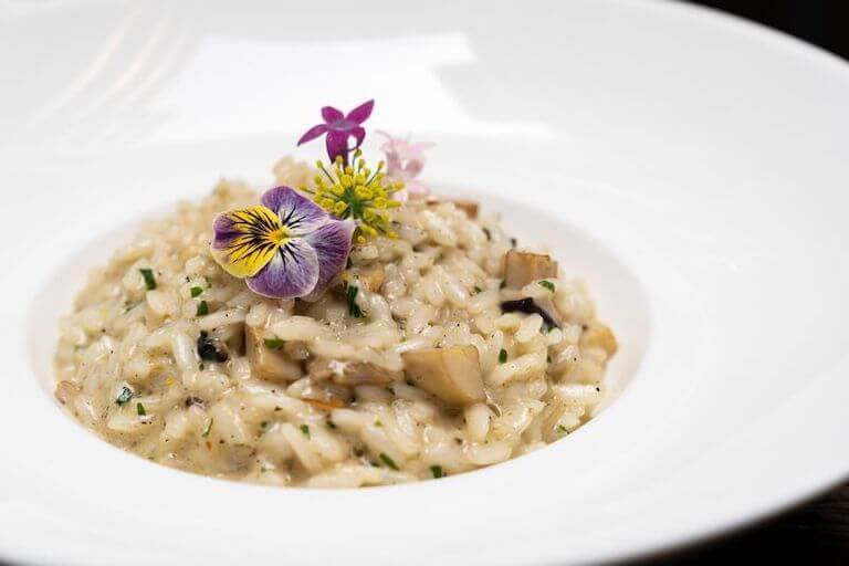 Mushroom risotto is plated on a white bowl, and topped with brightly colored pink and yellow flowers.