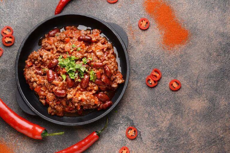 Cast iron dish of chili with beans on a table surrounded by whole and sliced chili peppers.