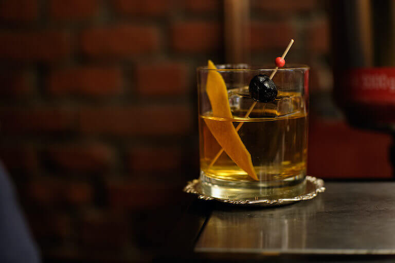 An upscale old fashioned cocktail sits precariously on the edge of a table.