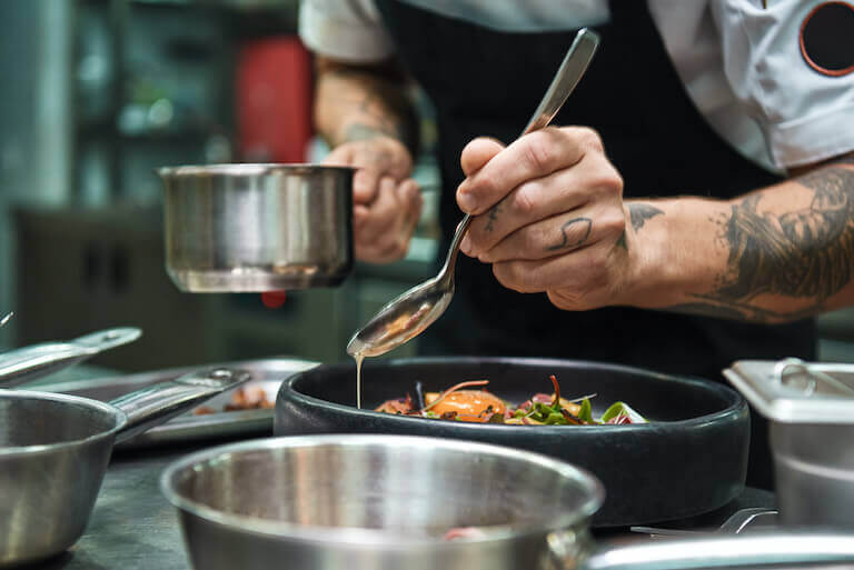 A cook with tattoos drizzles sauce over a fresh dish on a black plate.