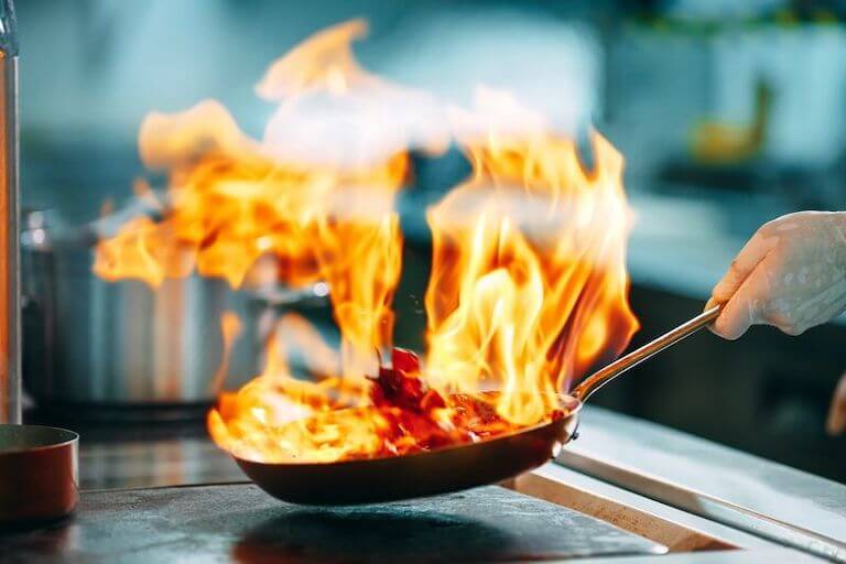 A hand shaking a pan above a stove top with large flames emanating from the cooking food.