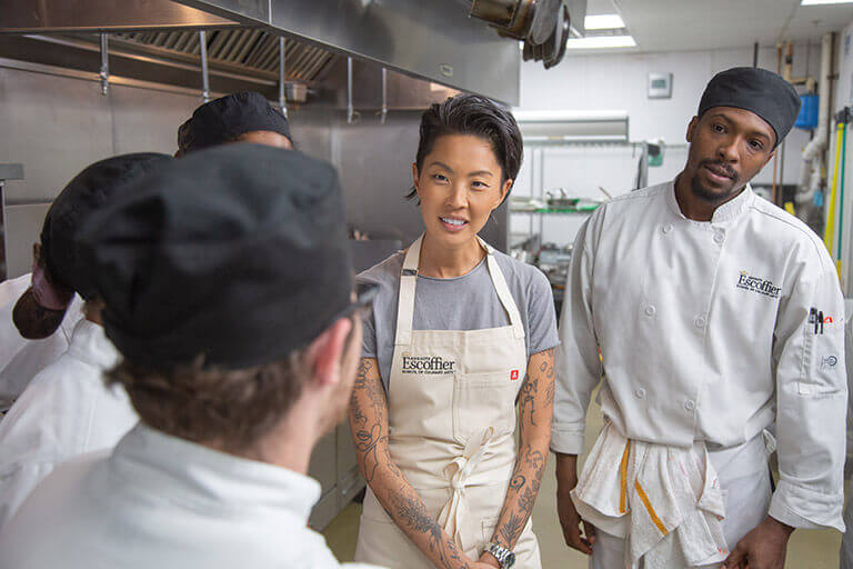 Chef Kristen Kish stands inside of a commercial kitchen wearing an Escoffier apron and chatting with several other peers. 