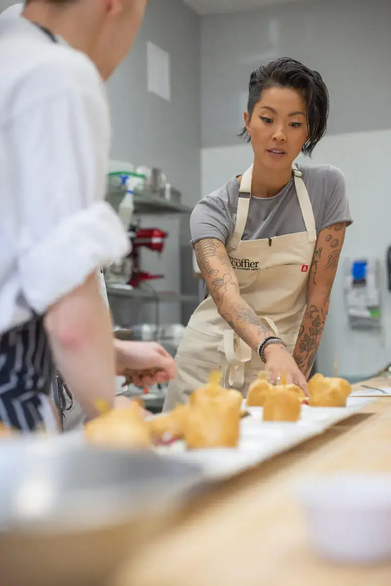 Kristen Kish wears a chef’s apron inside of a kitchen and points out details about baked goods to another person whose back is to the camera.